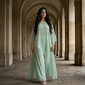A woman stands in a stone colonnade wearing a flowing mint green two-piece outfit with a loose long-sleeve top and wide-leg pants. She is wearing gold bracelets and sandals.