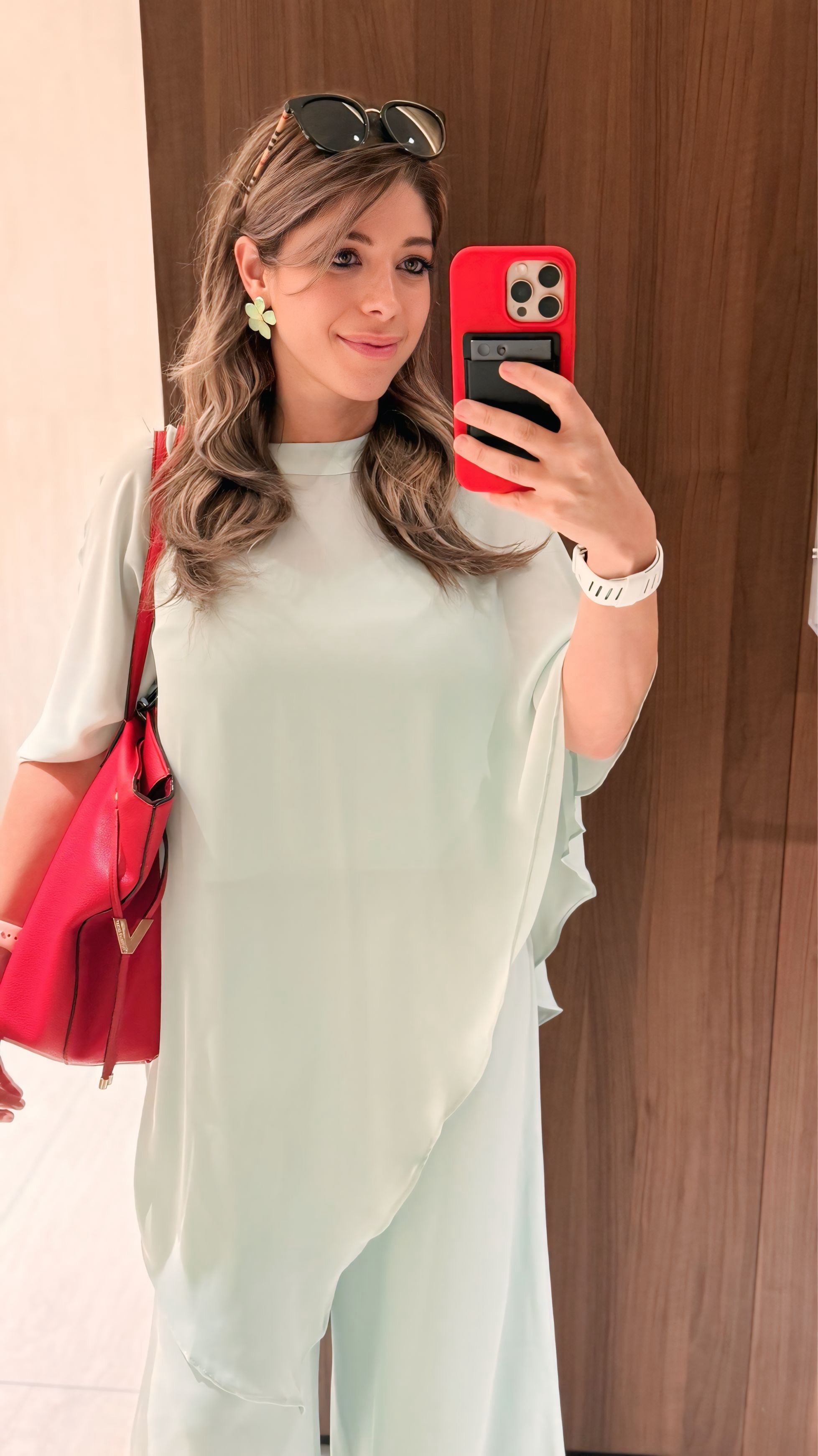 A woman taking a mirror selfie wearing a light sage green oversized tunic top and matching wide-leg trousers, carrying a bright red shoulder bag and holding a red smartphone.