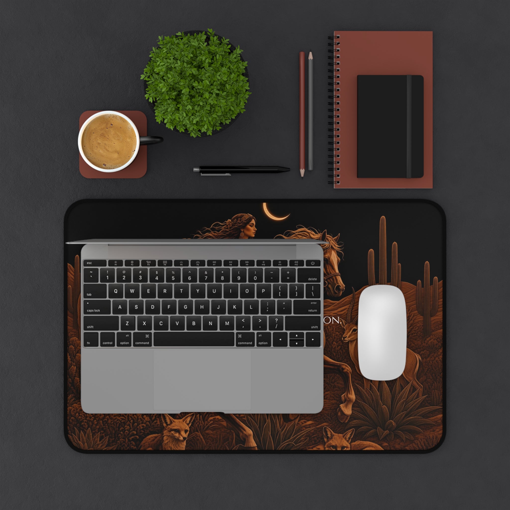 A top-down flat lay of a large desk mat with a desert-themed illustration, styled with a laptop, mouse, plant, coffee cup, and notebook on a dark gray surface.
